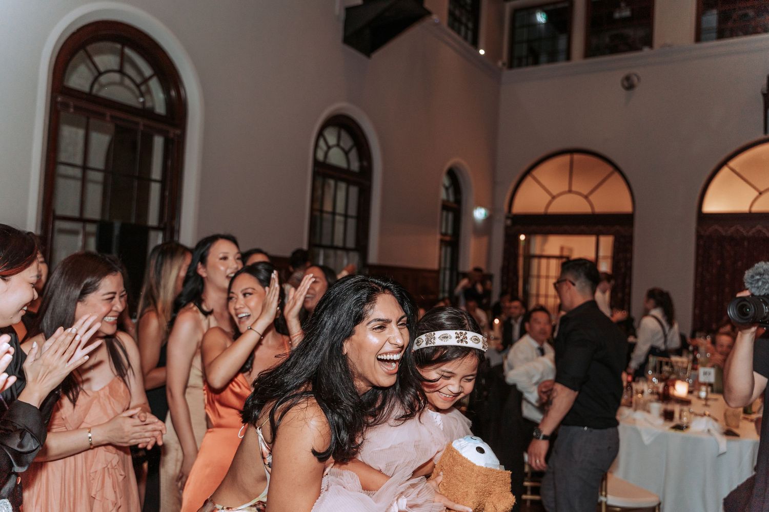 Enthusiastic wedding guests dancing and celebrating at reception venue with high ceilings.