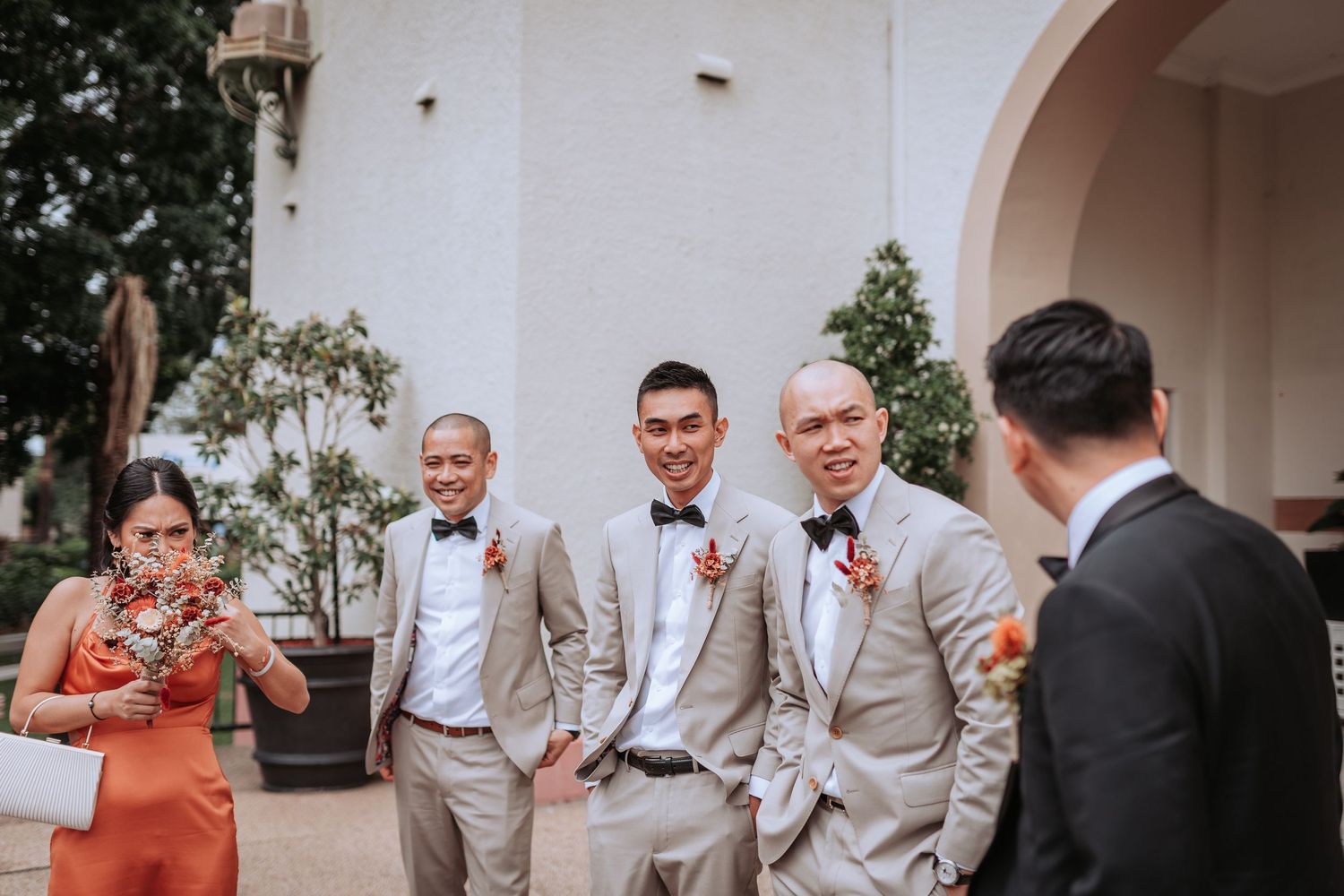 Wedding party members in light colored suits share drinks and laughter outdoors.