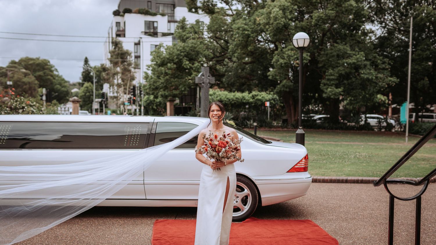 A bride with flowing veil stands on a red carpet beside a luxury car.