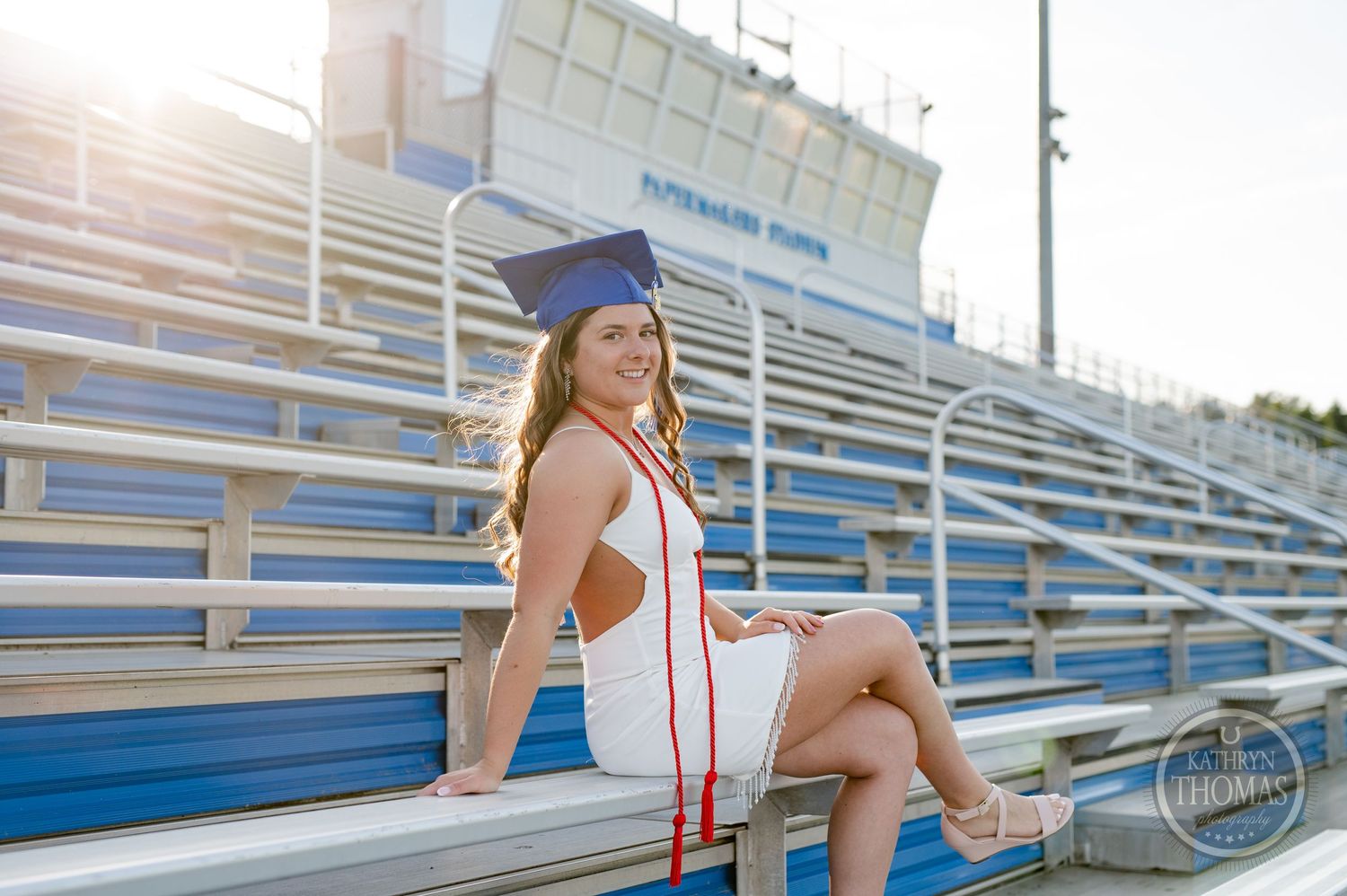 Graduate in white dress sits on stadium bleachers during sunset photo session.
