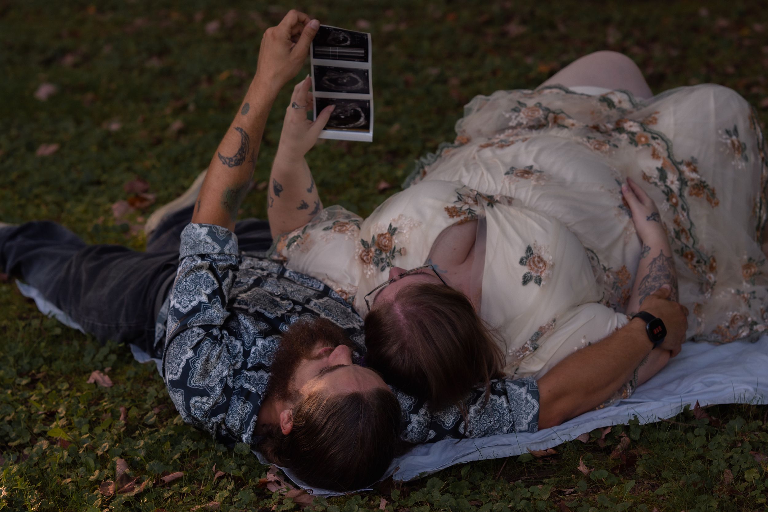 Couple lying on grass looking at ultrasound photos during outdoor maternity photoshoot at dusk.