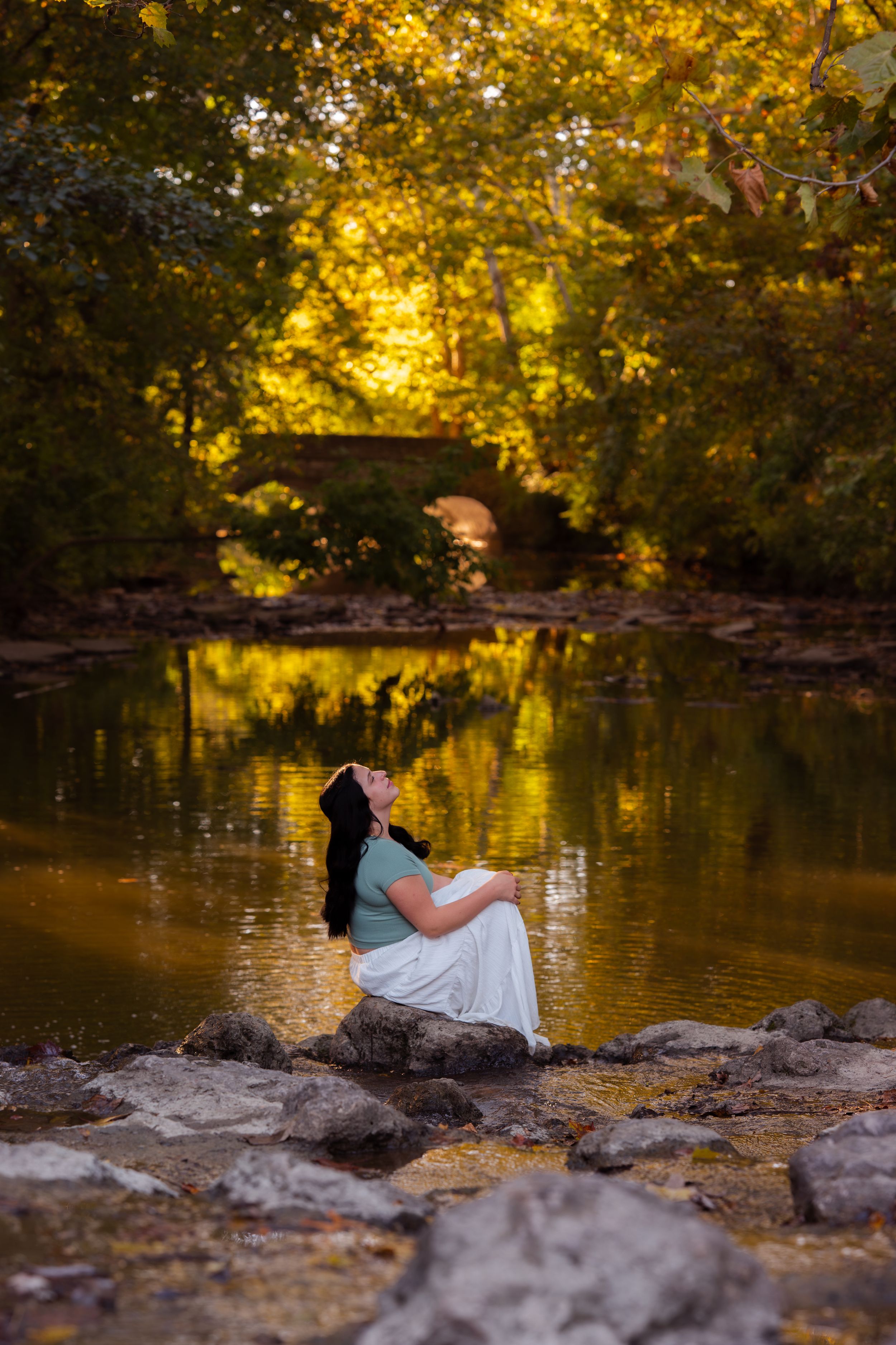 Pregnant woman in white skirt sits on rocks by a golden reflective pond surrounded by autumn trees.