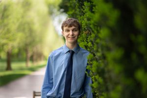 A student in formal attire poses outdoors on a tree-lined path with lush spring greenery in the background.