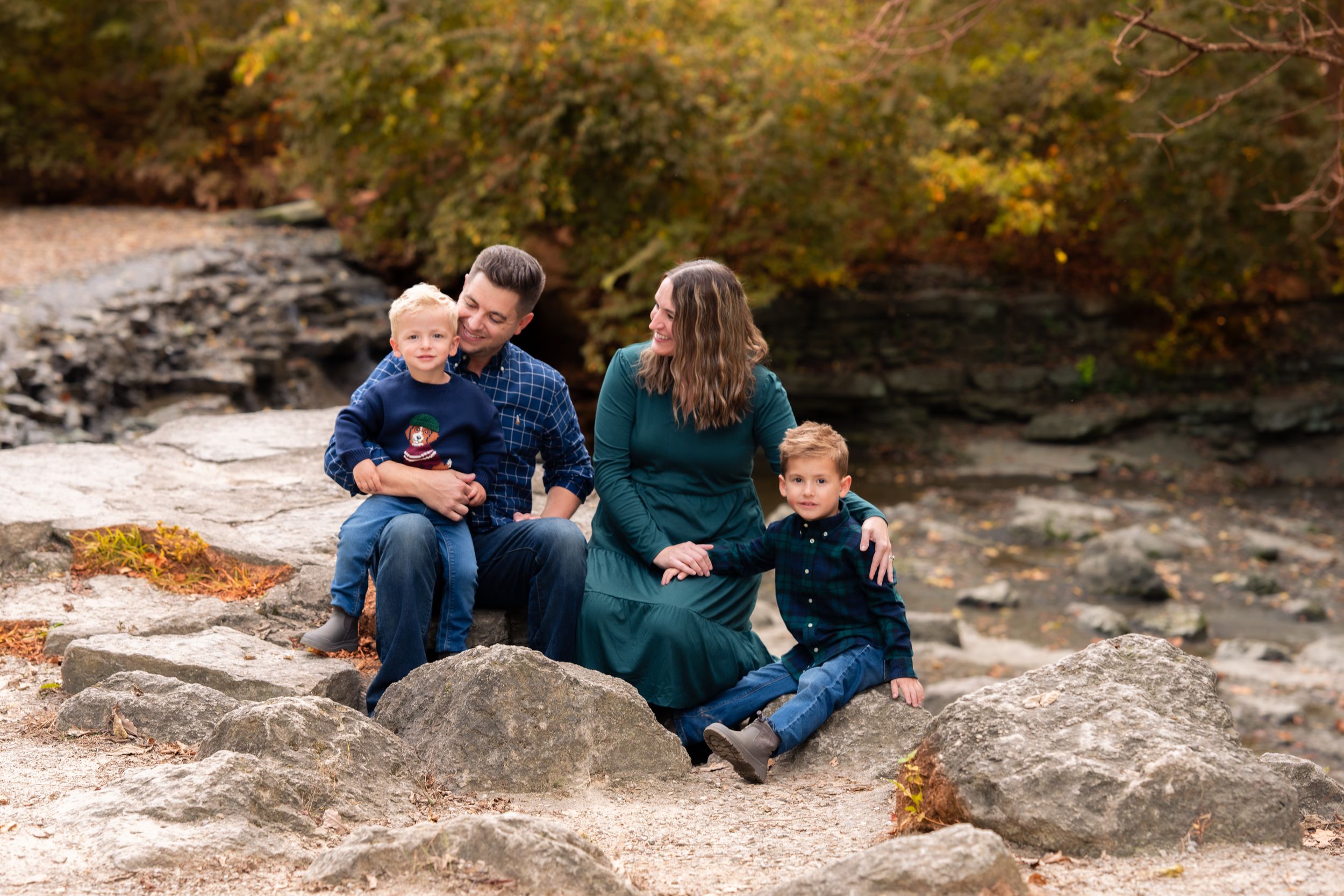 Family of four sitting on rocks outdoors in autumn, two young boys with parents in fall foliage setting.