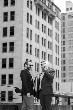Two men in business attire having a conversation on a rooftop with urban buildings in the background in black and white.