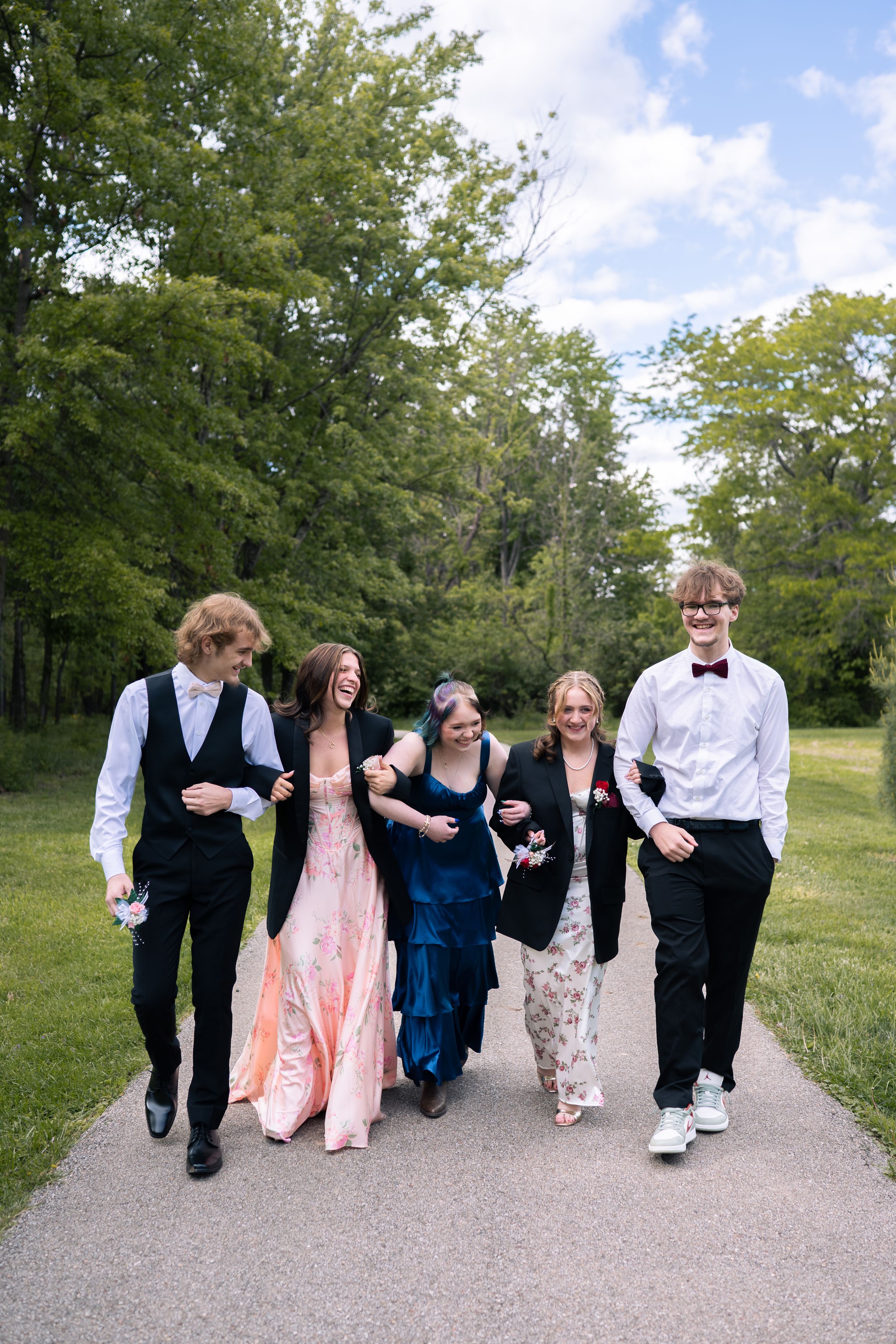 Five teenagers in formal prom attire walk and laugh together on a path surrounded by lush green trees.