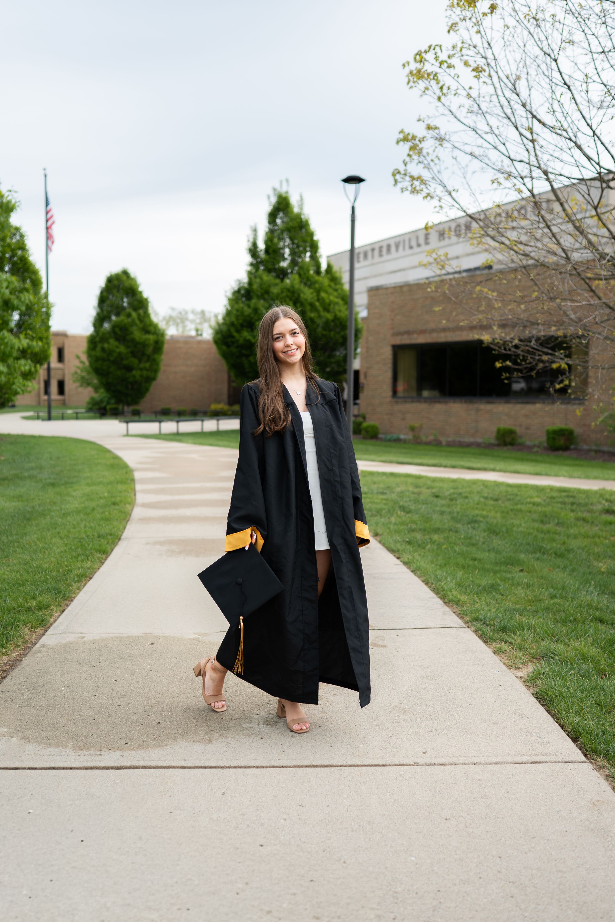 Young woman in black graduation gown and cap smiling on a school sidewalk on a cloudy spring day.