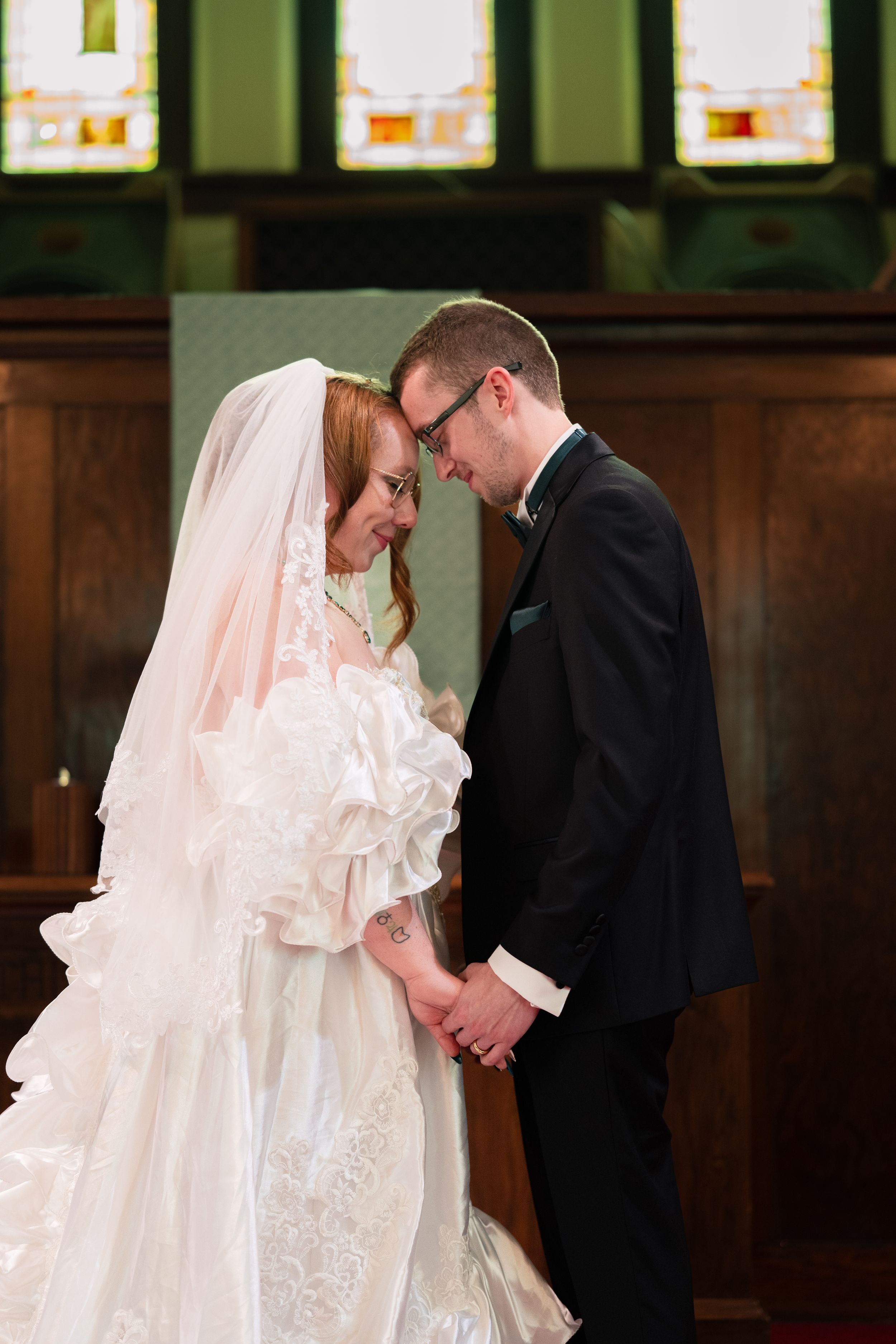 Bride and groom hold hands and touch foreheads in a church, bride wearing white lace gown with veil, groom in black tuxedo.