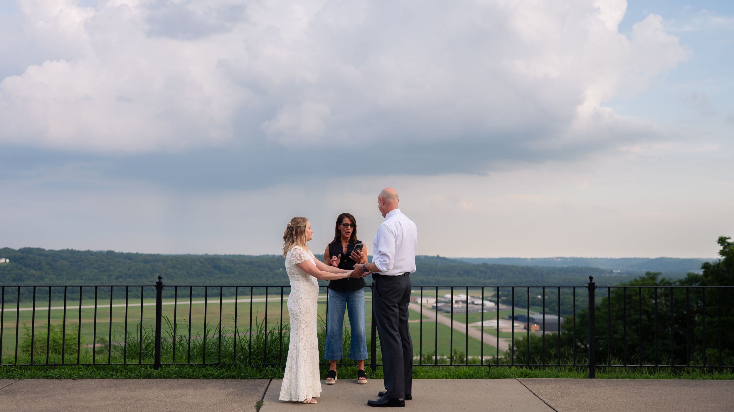Couple exchanges vows with officiant at scenic overlook with lush green valley and dramatic cloudy sky backdrop.