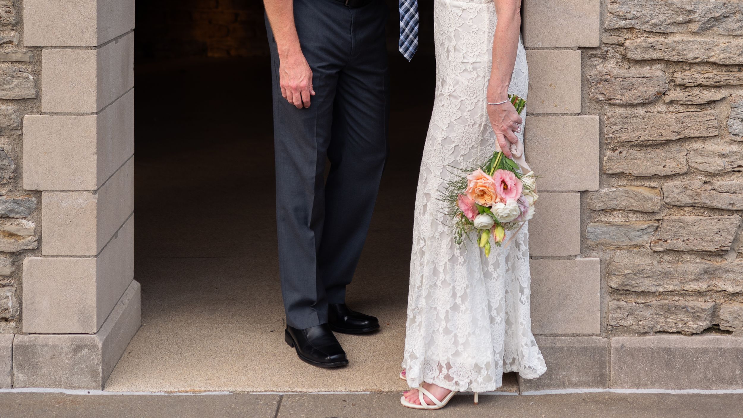 Bride in lace gown holding colorful bouquet stands with groom in navy suit at stone church entrance.