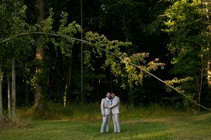 A couple embraces while standing in a wooded area with sunlight filtering through green trees.