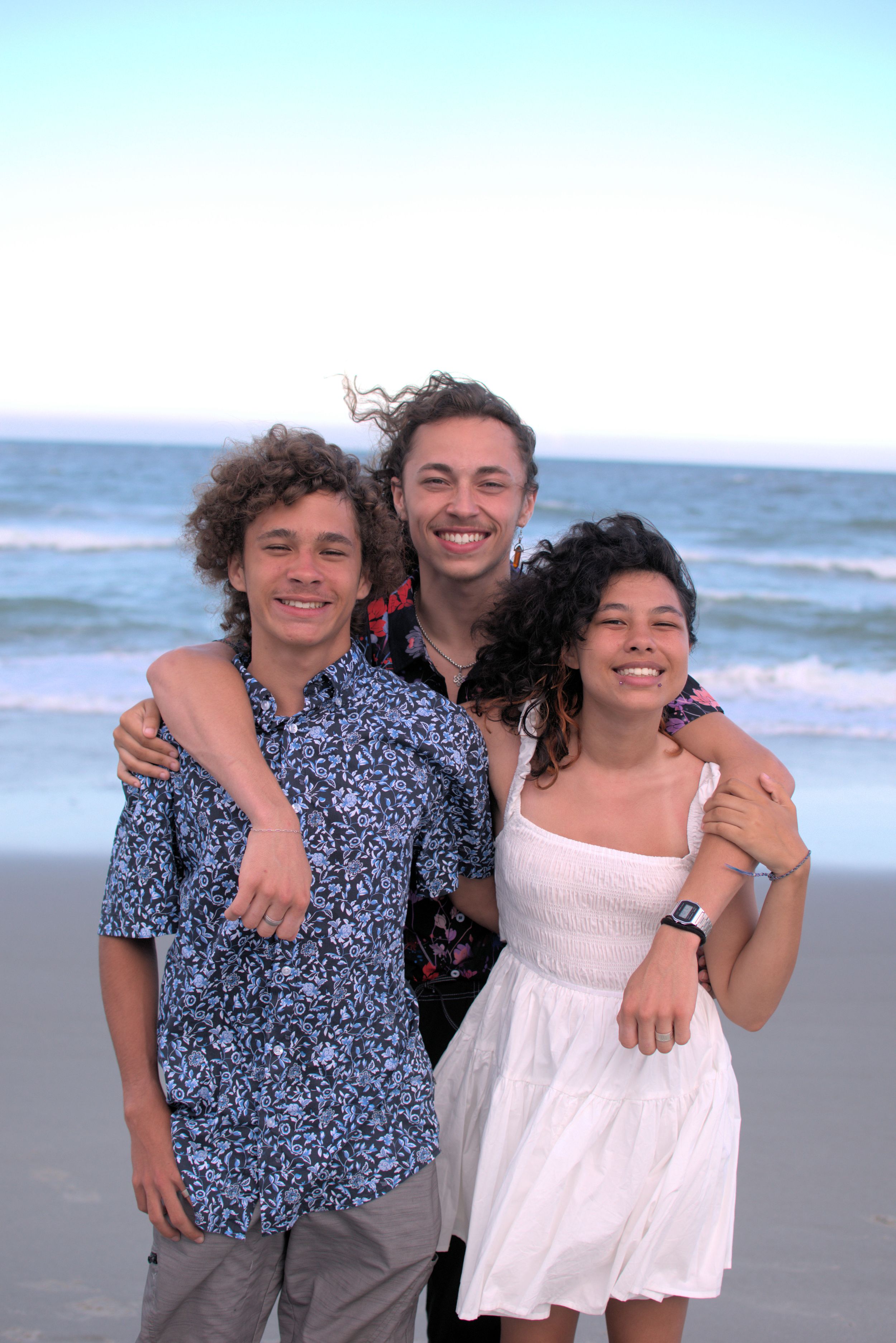 Three companions share a joyful moment together on a sunny beach with ocean waves in the background.