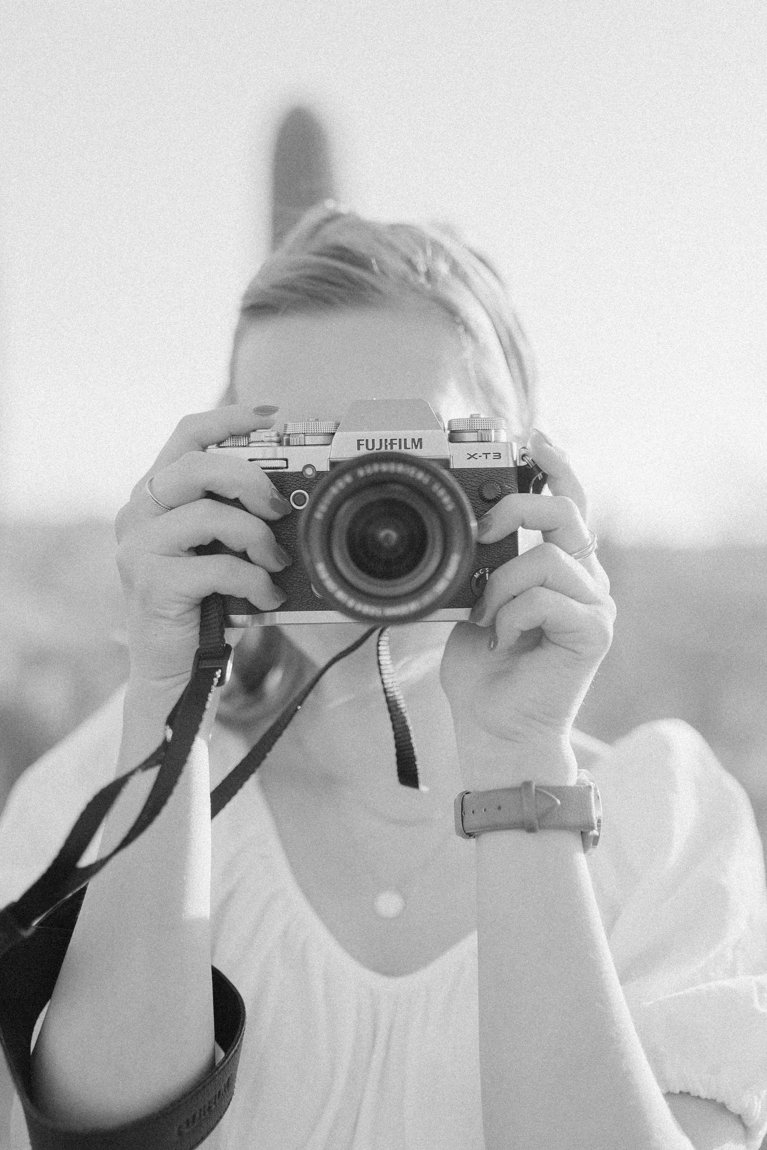 A person holds up a Fujifilm mirrorless camera to take a photograph in a black and white image.