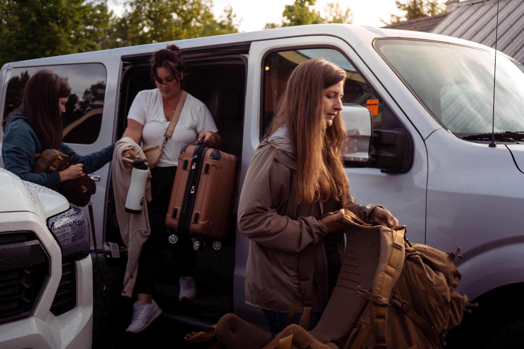 Group of travelers loading backpacks and luggage into a silver van on a sunny day.