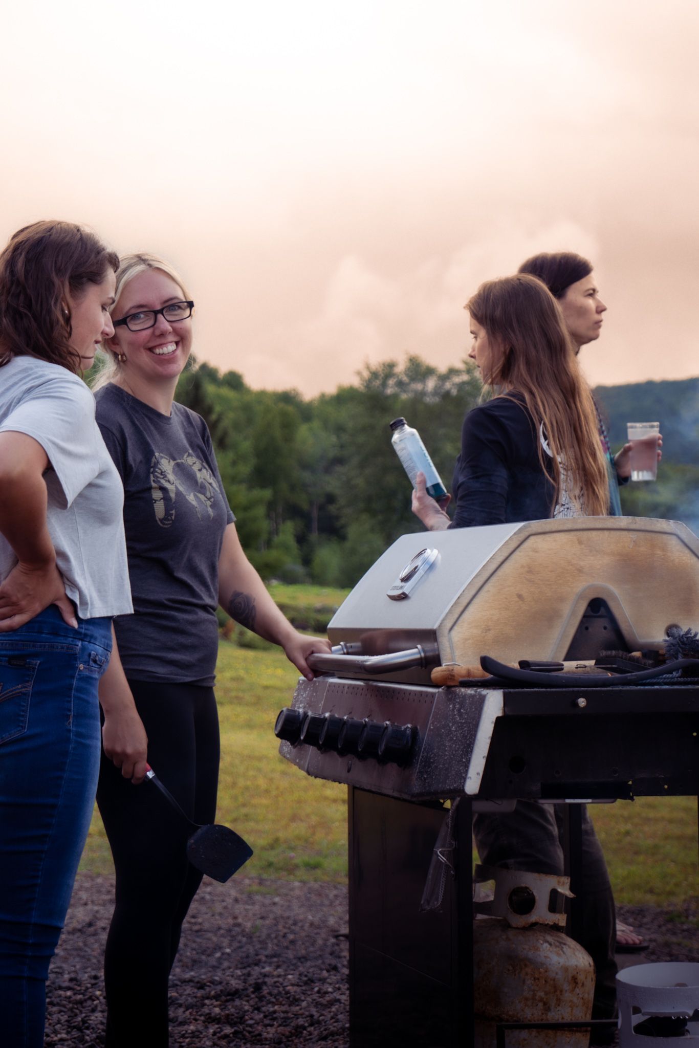 A group of people gather around a barbecue grill at sunset in an outdoor setting with trees in the background.
