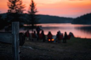 A group of silhouetted figures gather around a campfire by a serene lake at sunset with pine trees in the background.