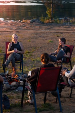 A group sits in camping chairs around a campfire by the water at sunset enjoying an outdoor gathering.