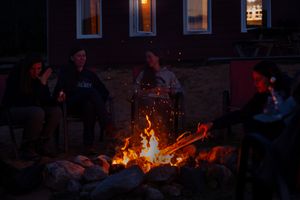 People gather around a glowing campfire at night outside a house with lit windows in the background.