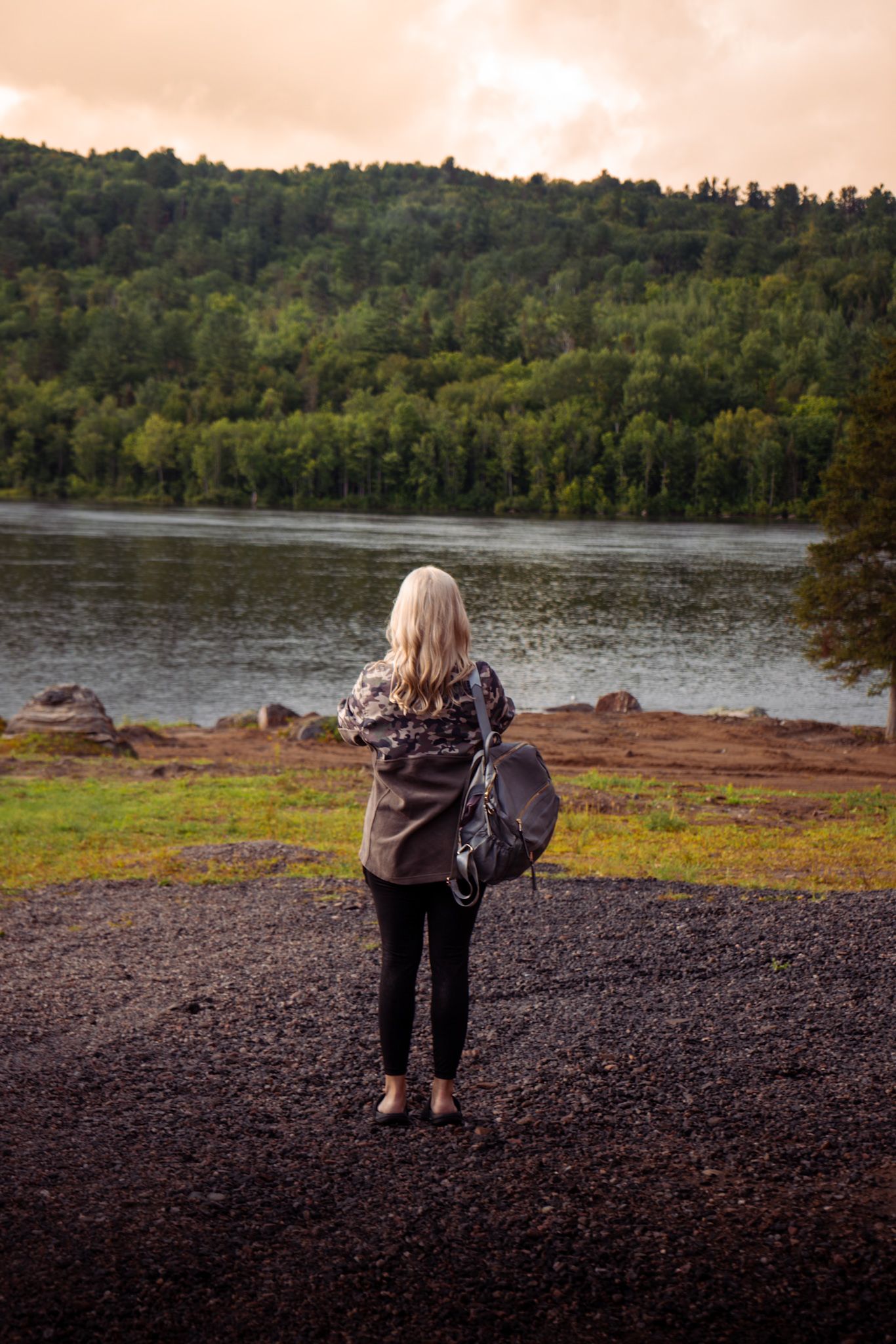 Person in casual wear with backpack stands on rocky shoreline overlooking serene lake surrounded by forested hills at sunset.