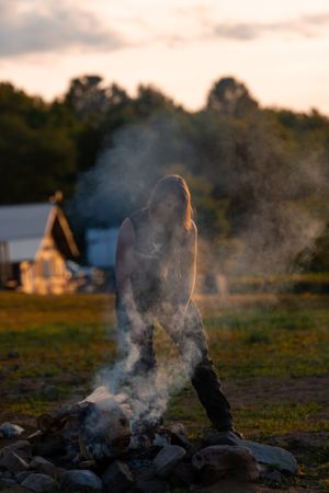 Silhouette of a person standing near a campfire with smoke rising at sunset with a cabin in the background.
