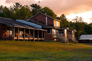 A rustic cabin lodge with a covered porch sits on a grassy hillside at sunset with pine trees in the background.