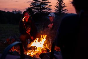 Friends gather around a glowing campfire in the woods at dusk with pine trees silhouetted against an orange sunset sky.