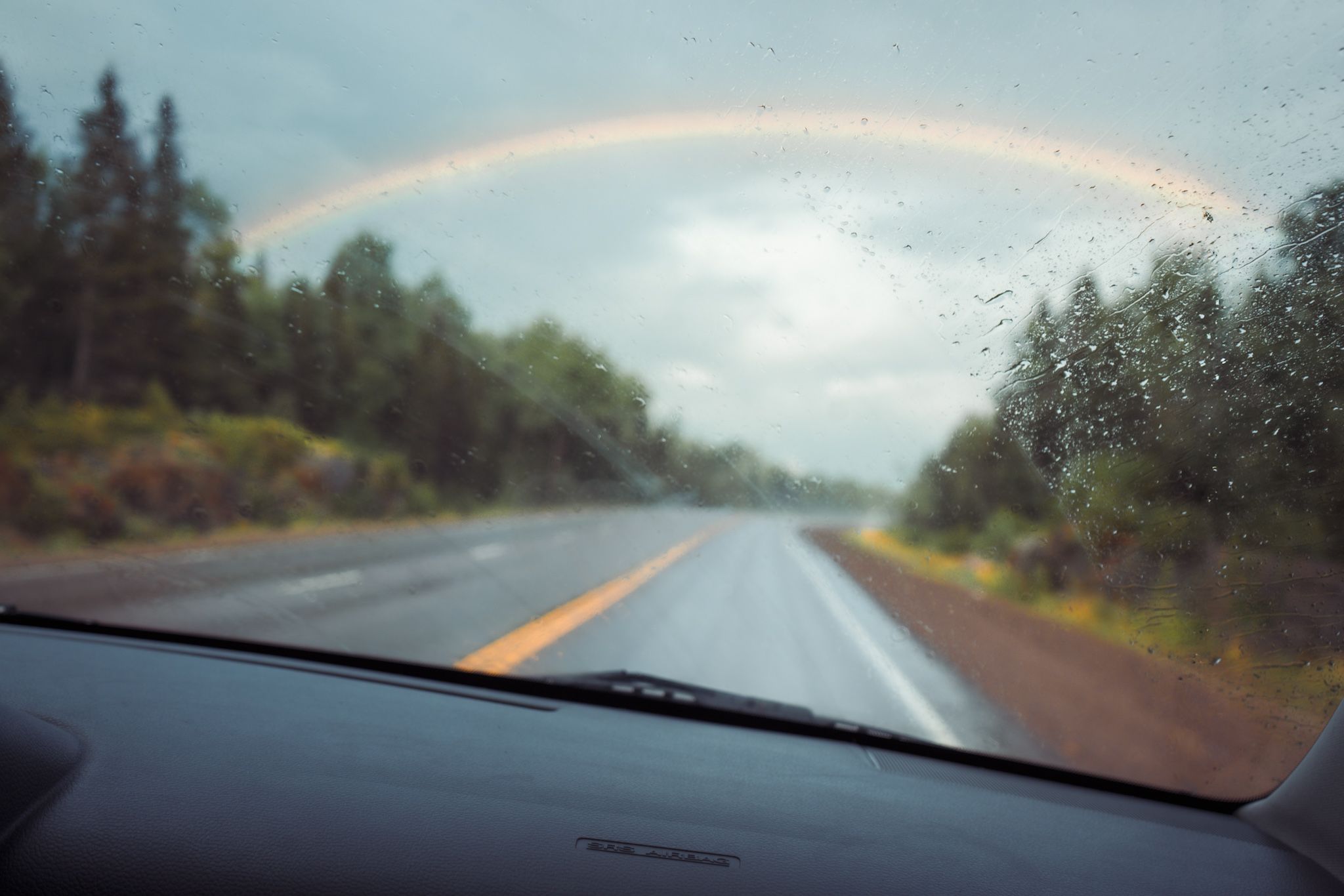 A colorful rainbow arches over a wet rural highway viewed through a car windshield on a rainy day.
