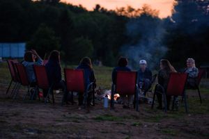 Group gathered around campfire in red lawn chairs at dusk enjoying an evening outdoors.
