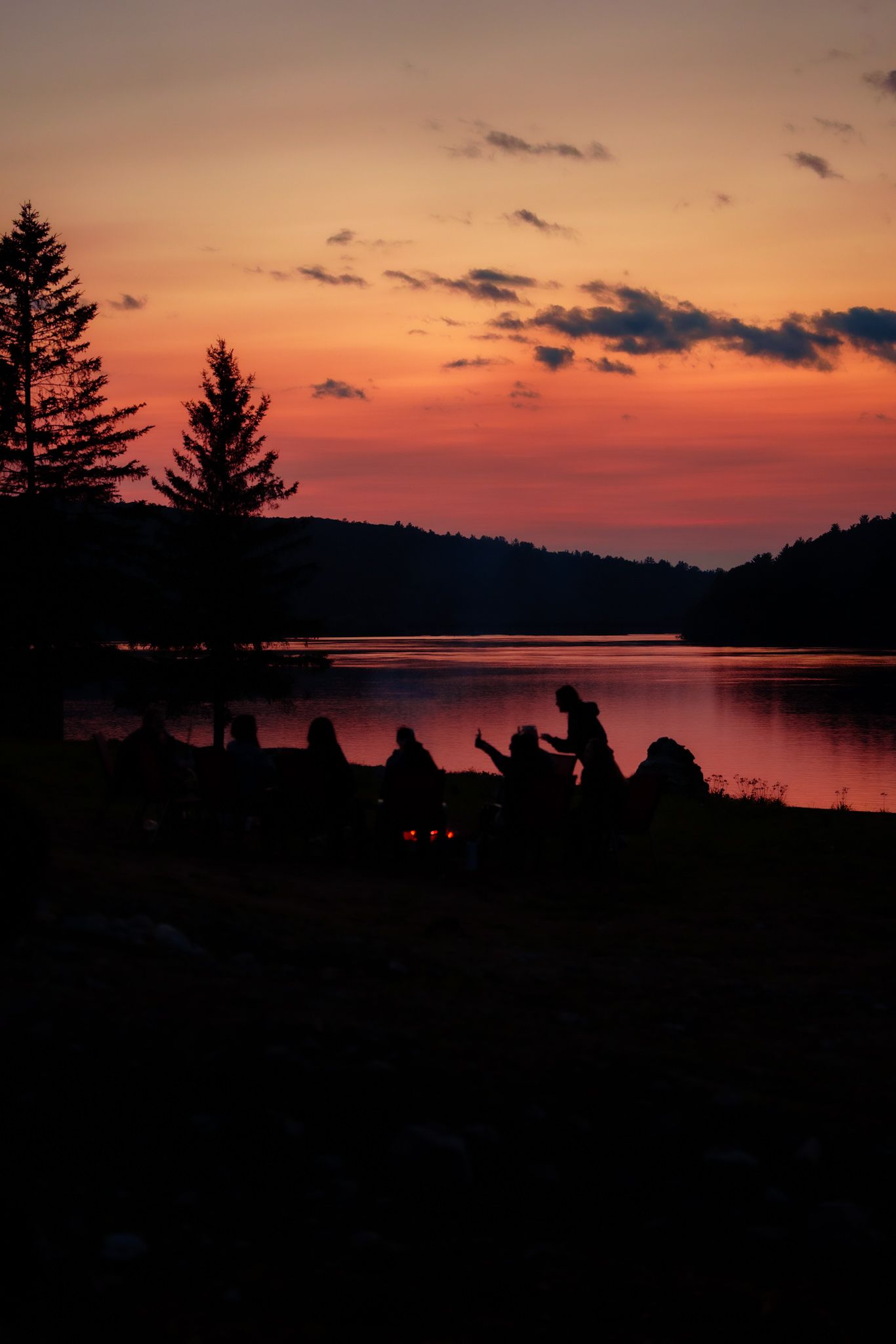 Silhouetted figures gathered by a tranquil lake at sunset with evergreen trees and dramatic orange sky reflected in the water.