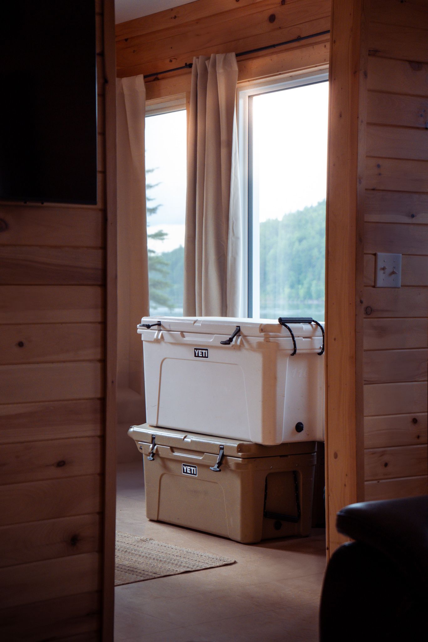 Two stacked YETI coolers sit near a window inside a wooden cabin interior with natural lighting and curtains.