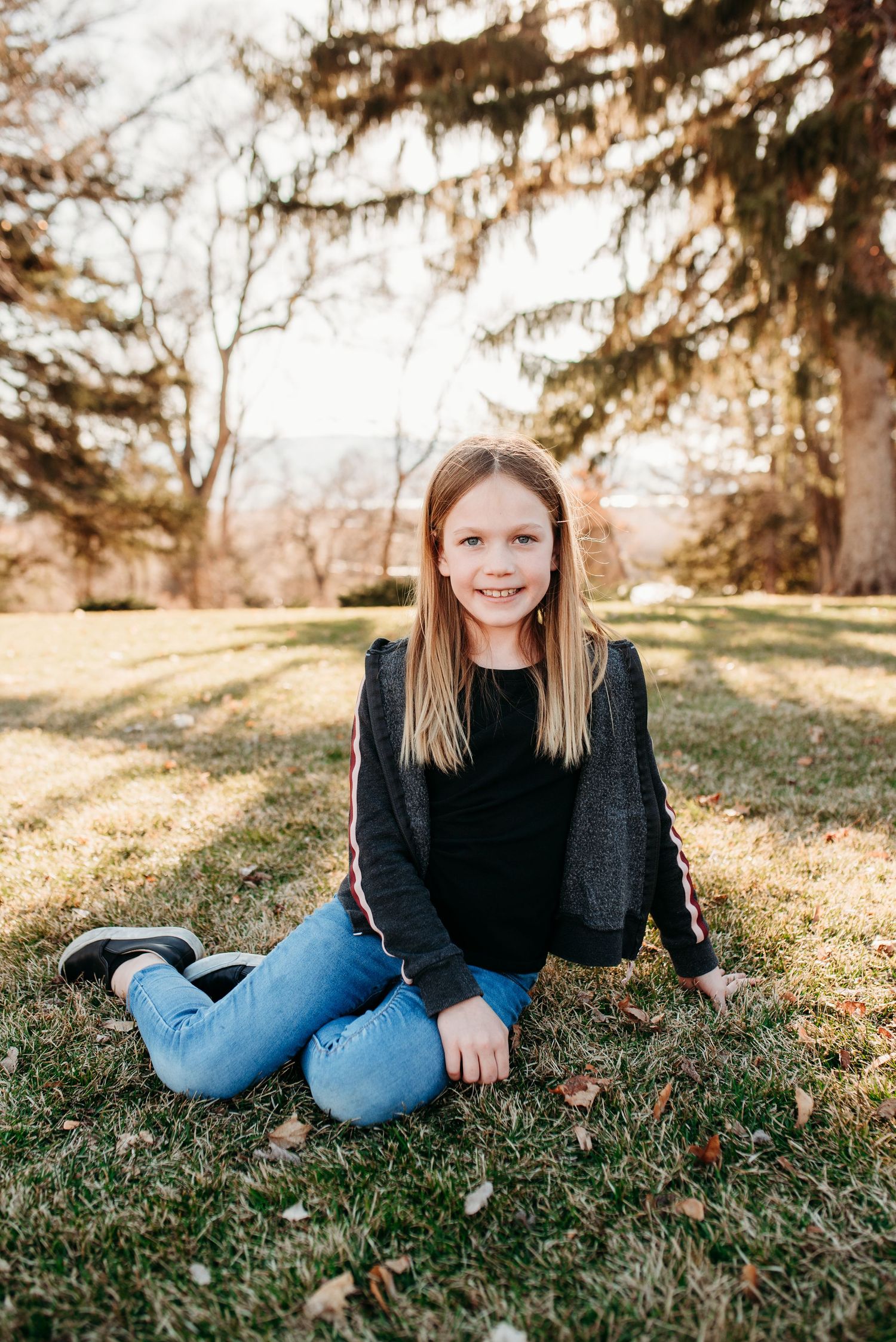 Young person in black sweater and jeans sitting on autumn grass with evergreen trees behind.
