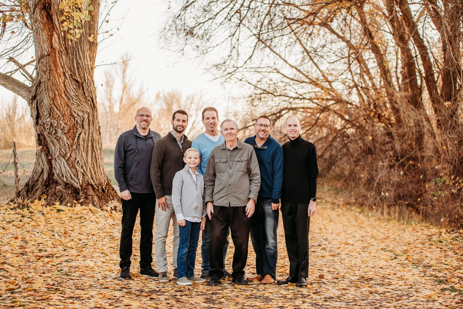 Group family photo taken outdoors with large trees and fallen autumn leaves.
