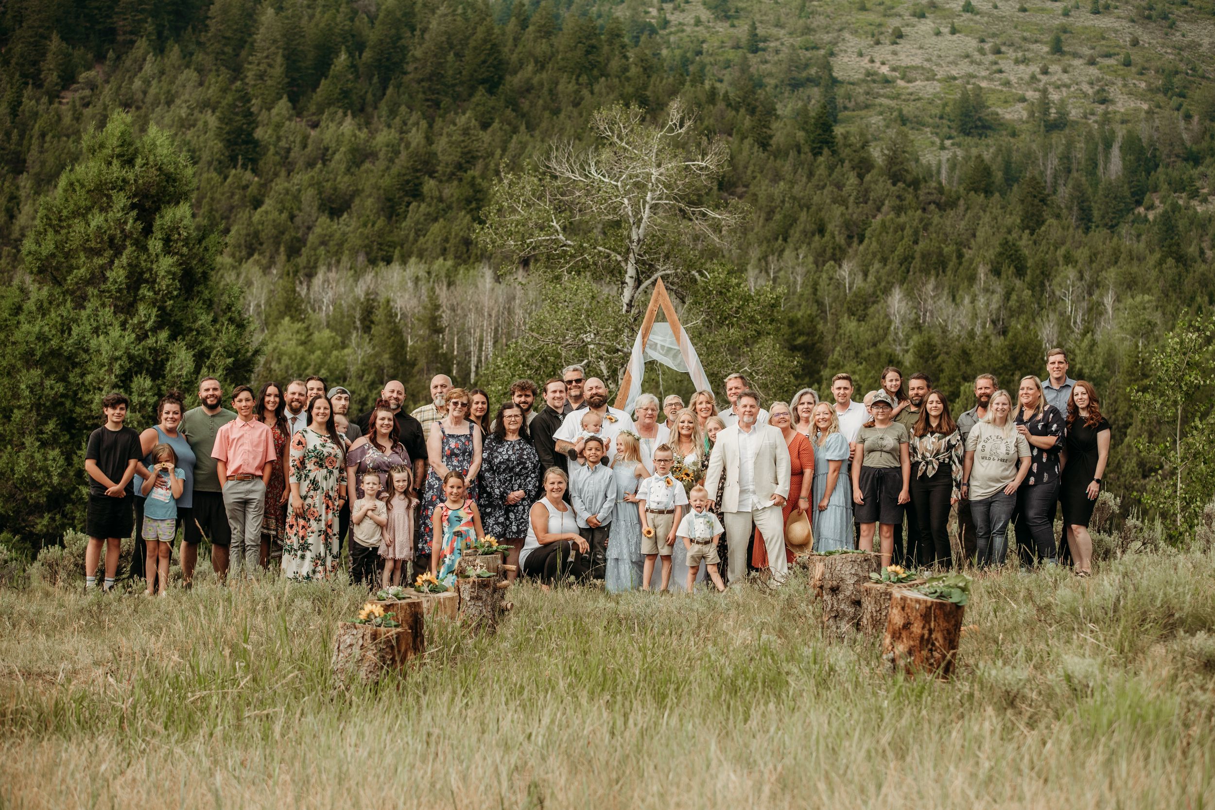 Large group photo of wedding guests gathered outdoors on a hillside with mountains in the background.