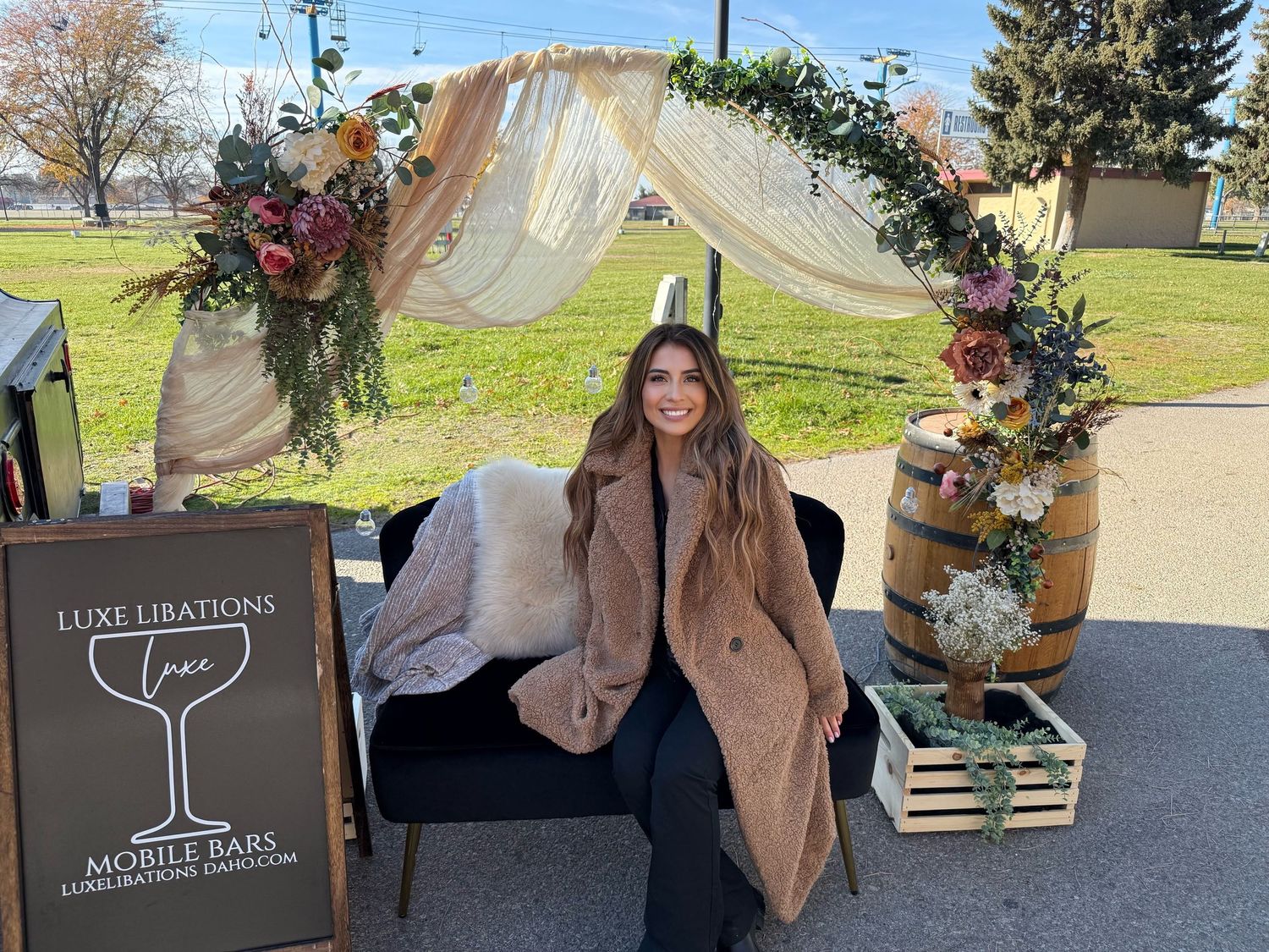 A decorated wedding arch with floral arrangements and white fabric stands outdoors next to a wine bar sign and wooden crates.