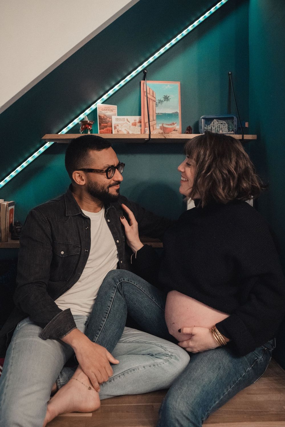 A couple sits together on the floor, smiling at each other in a cozy teal room with LED-lit shelves.