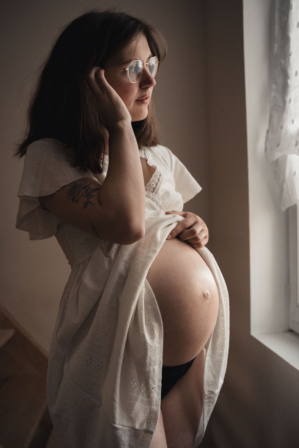 Pregnant woman in white robe and glasses gazes out window, cradling her baby bump in a softly lit room.