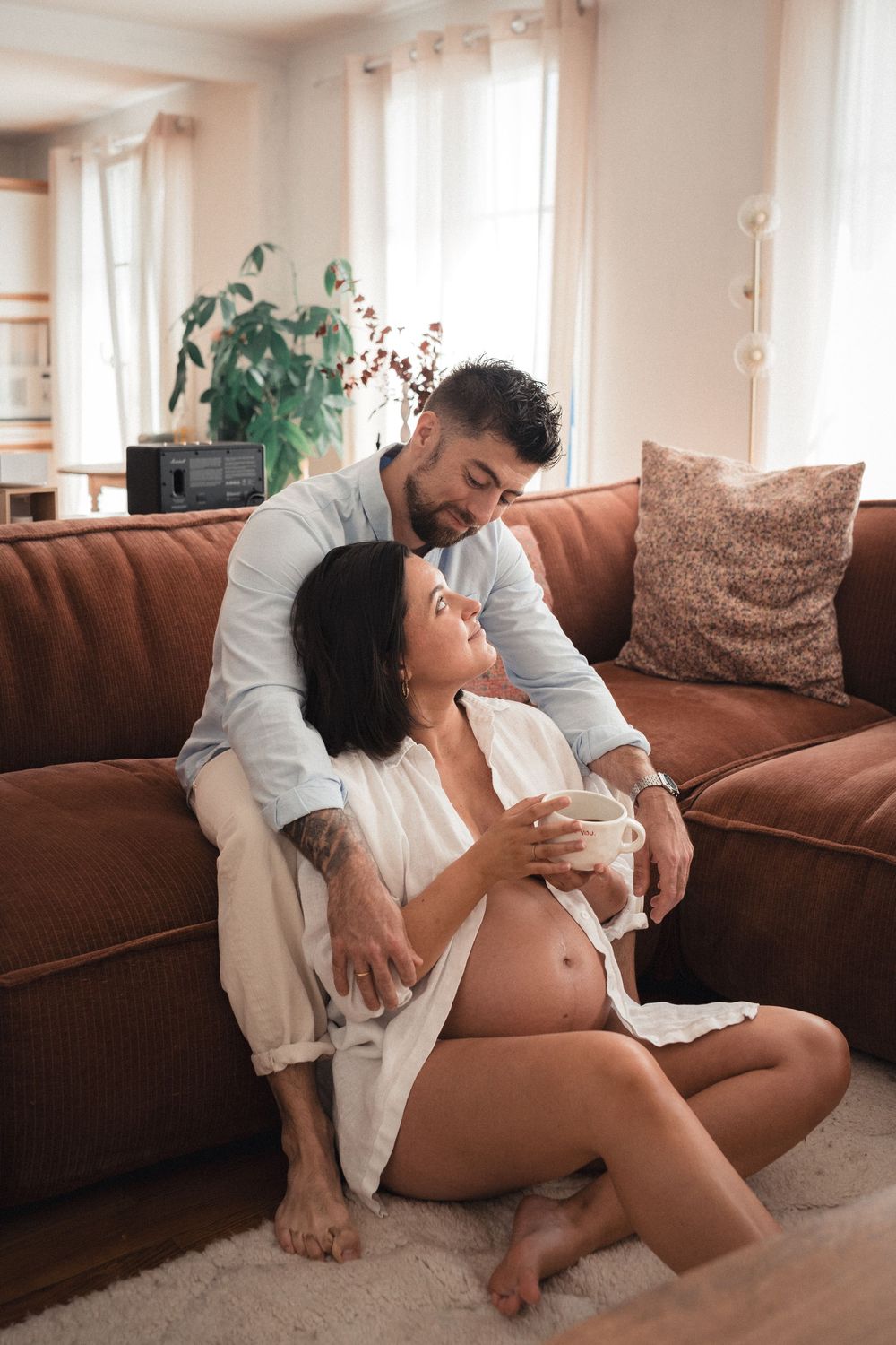 A couple relaxes together on a brown couch in a bright, sunlit living room.
