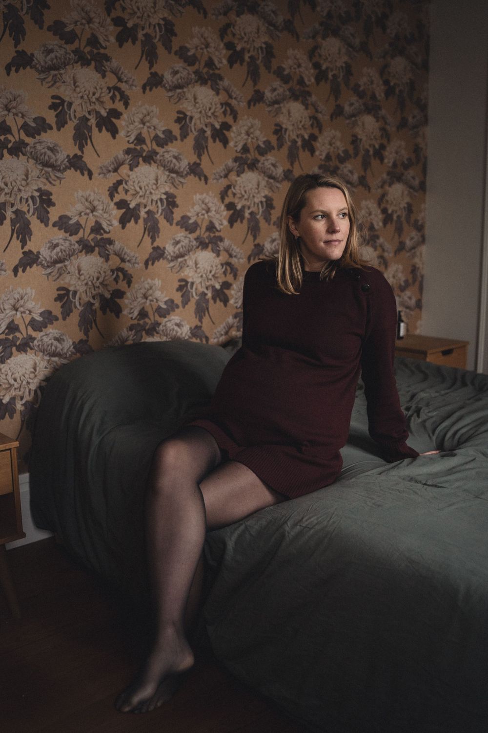 Femme enceinte en robe bordeaux assise sur un lit, avec en arrière-plan un papier peint fleuri.