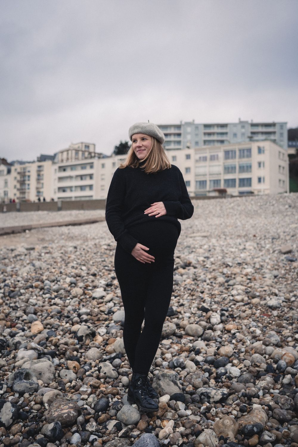 Femme enceinte se tenant le ventre rond en tenue d’hiver noire debout sur une plage de galets, avec des immeubles du Havre en arrière-plan.
