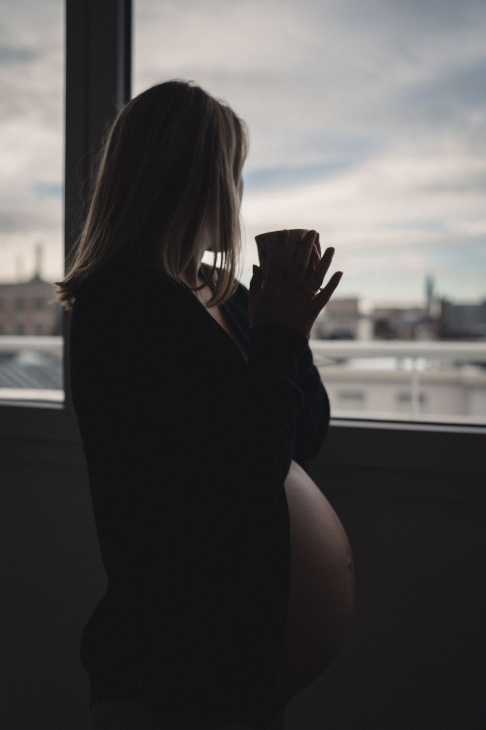 Silhouette of pregnant woman drinking from mug by window during sunset.