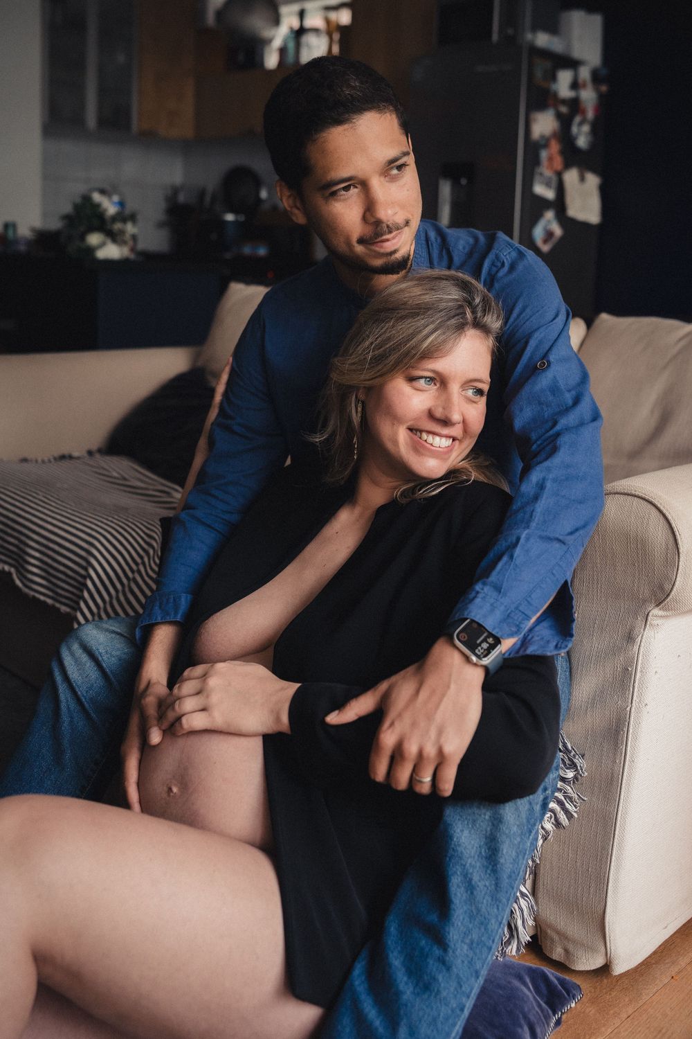 Intimate maternity photo series showing couple relaxing together on beige couch in dark room.