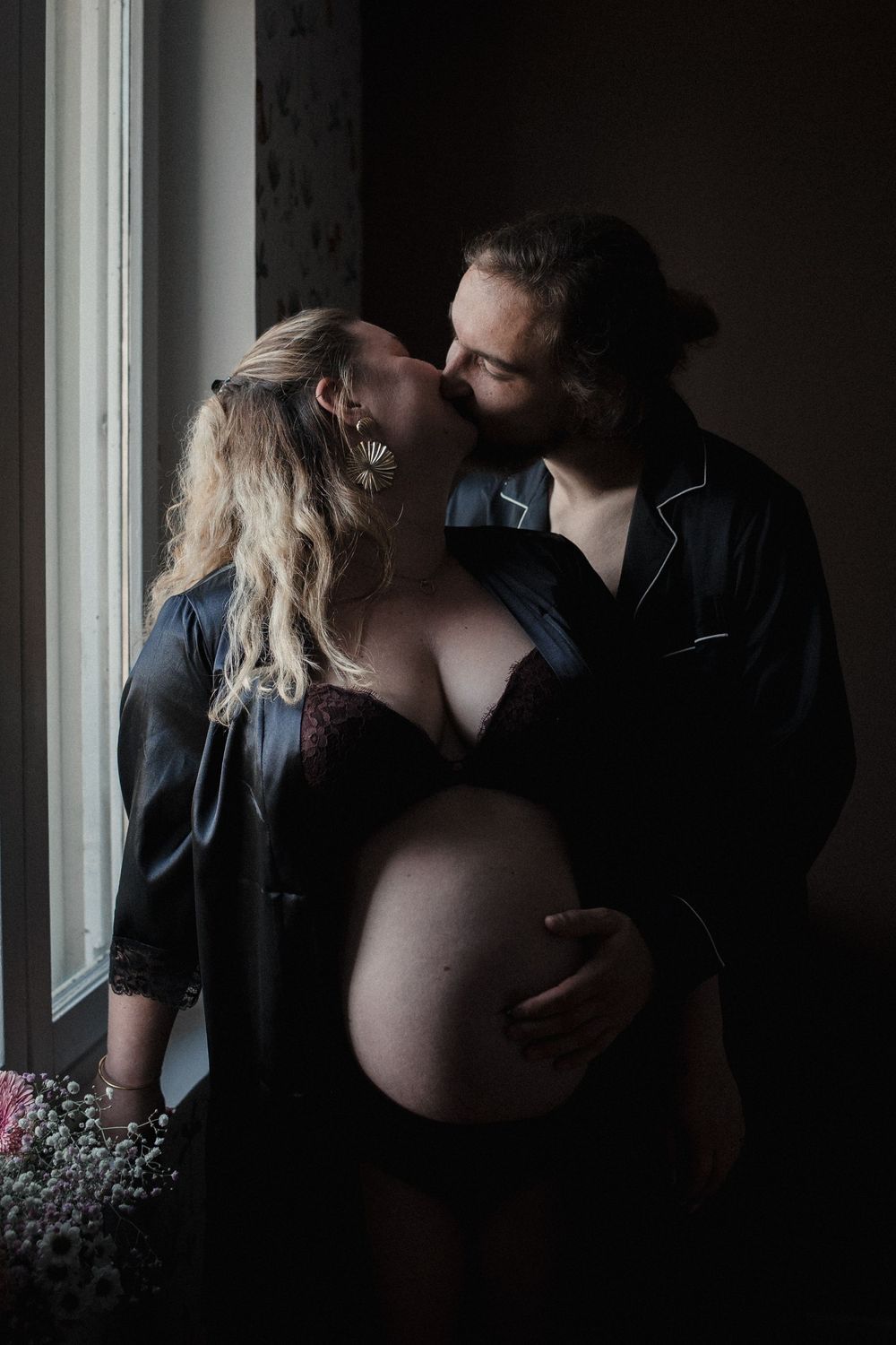 Silhouetted couple shares intimate moments in moody black and white maternity photoshoot by window.