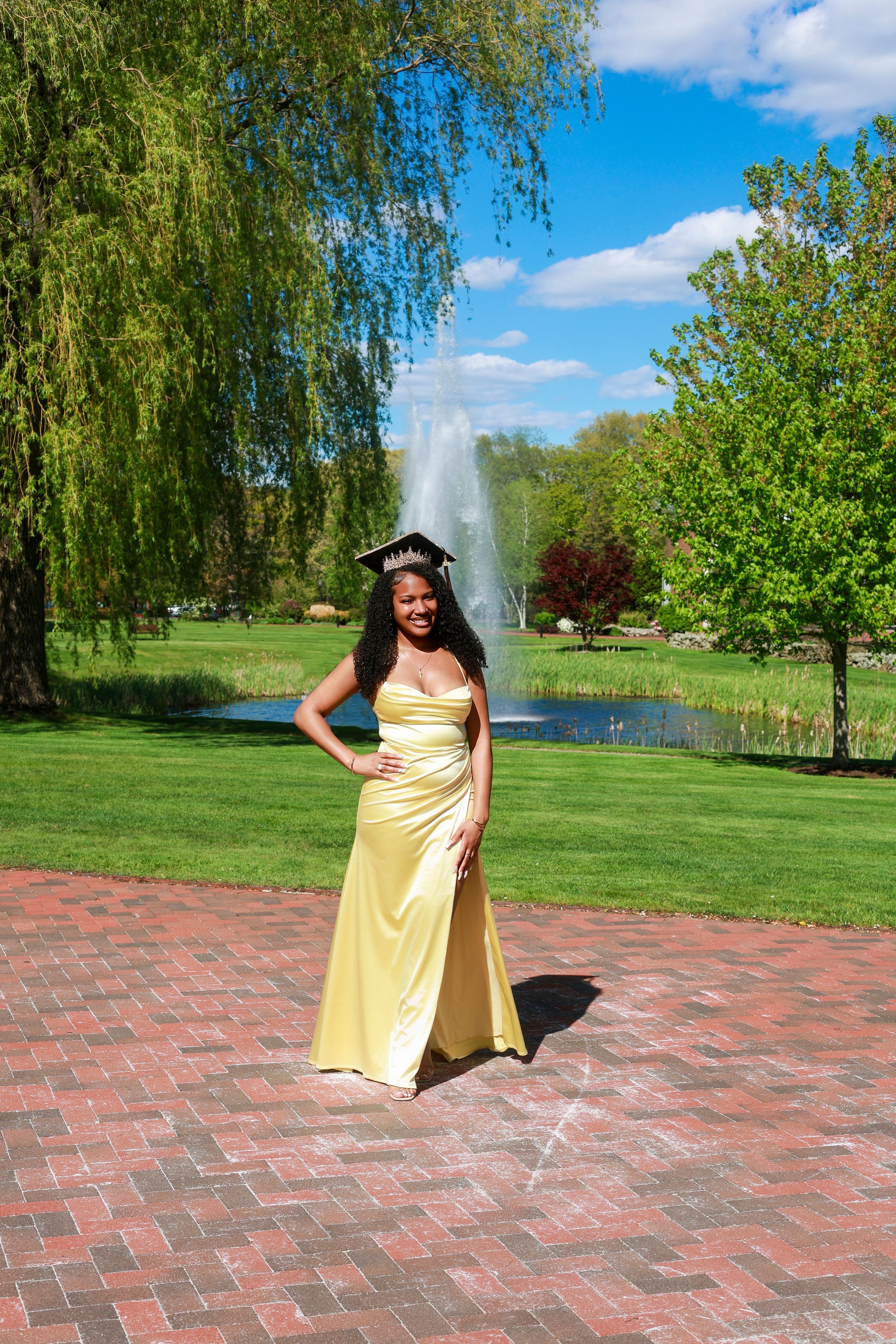 Young woman in yellow satin gown and graduation cap poses on brick path by fountain in lush green park.