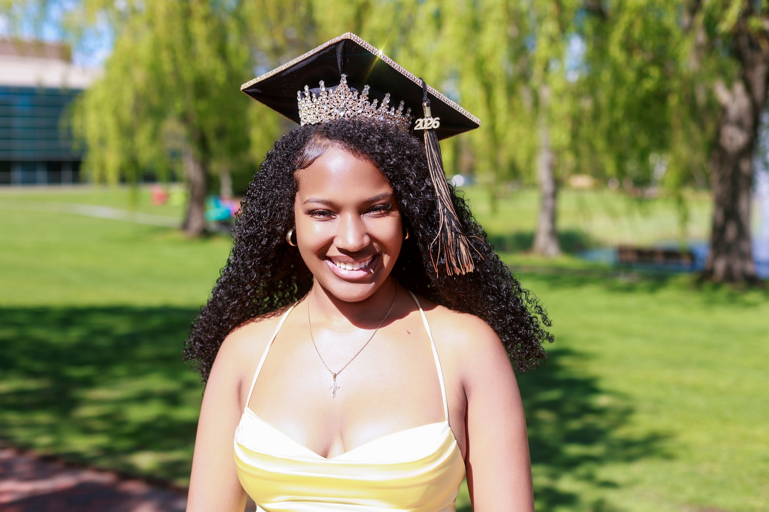 Smiling graduate wearing cap with tiara and yellow dress outdoors on sunny day.