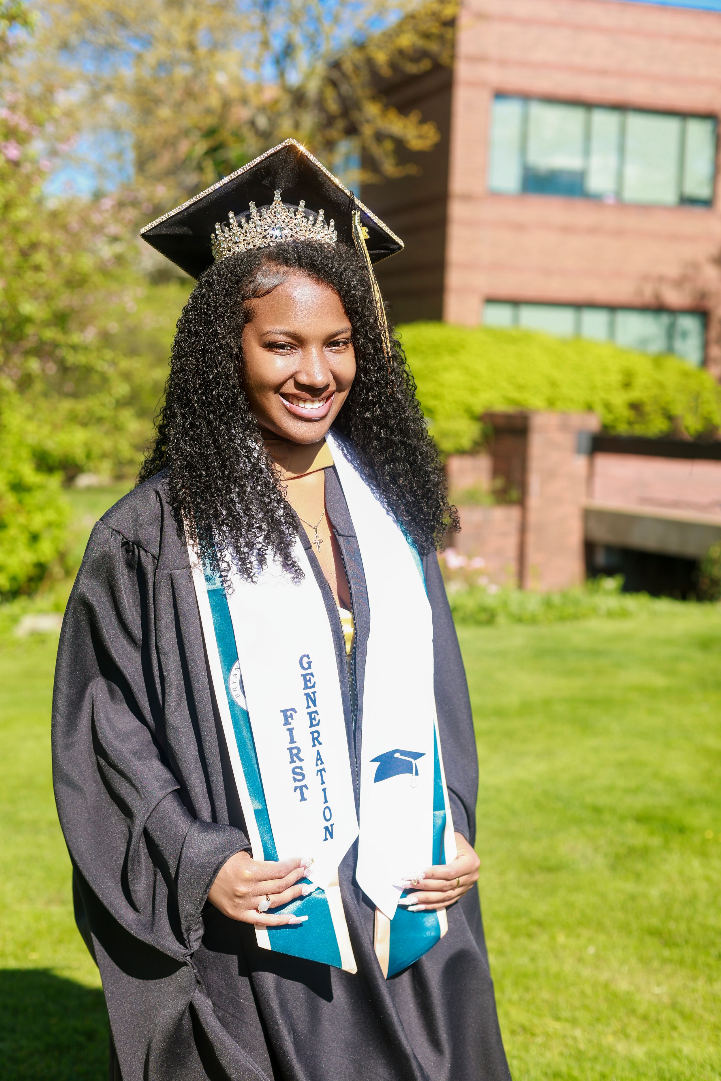 Smiling female graduate in cap, gown, tiara, and first generation stole poses outdoors on sunny campus.
