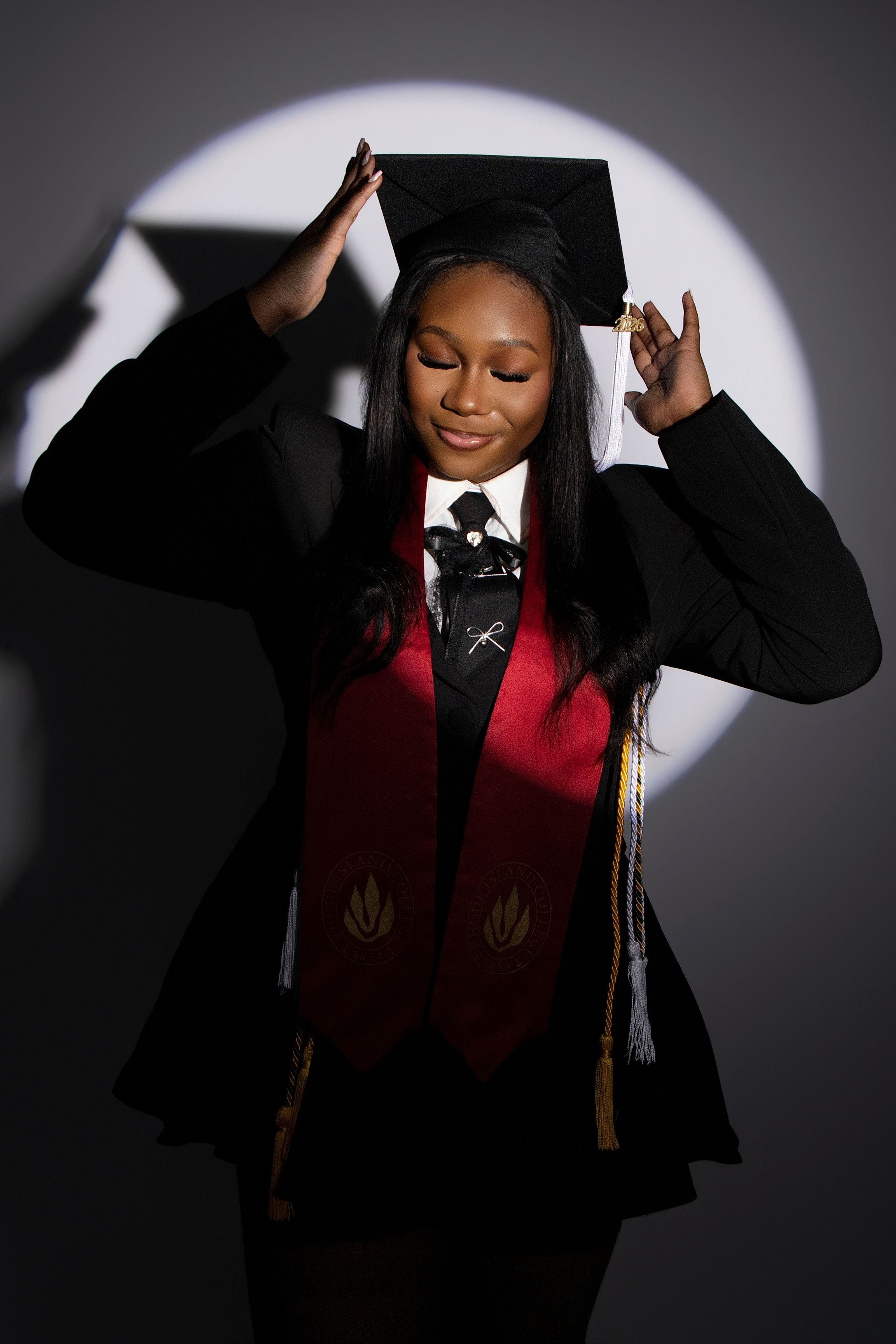 Young woman in graduation cap and gown with red stole smiles proudly under spotlight.