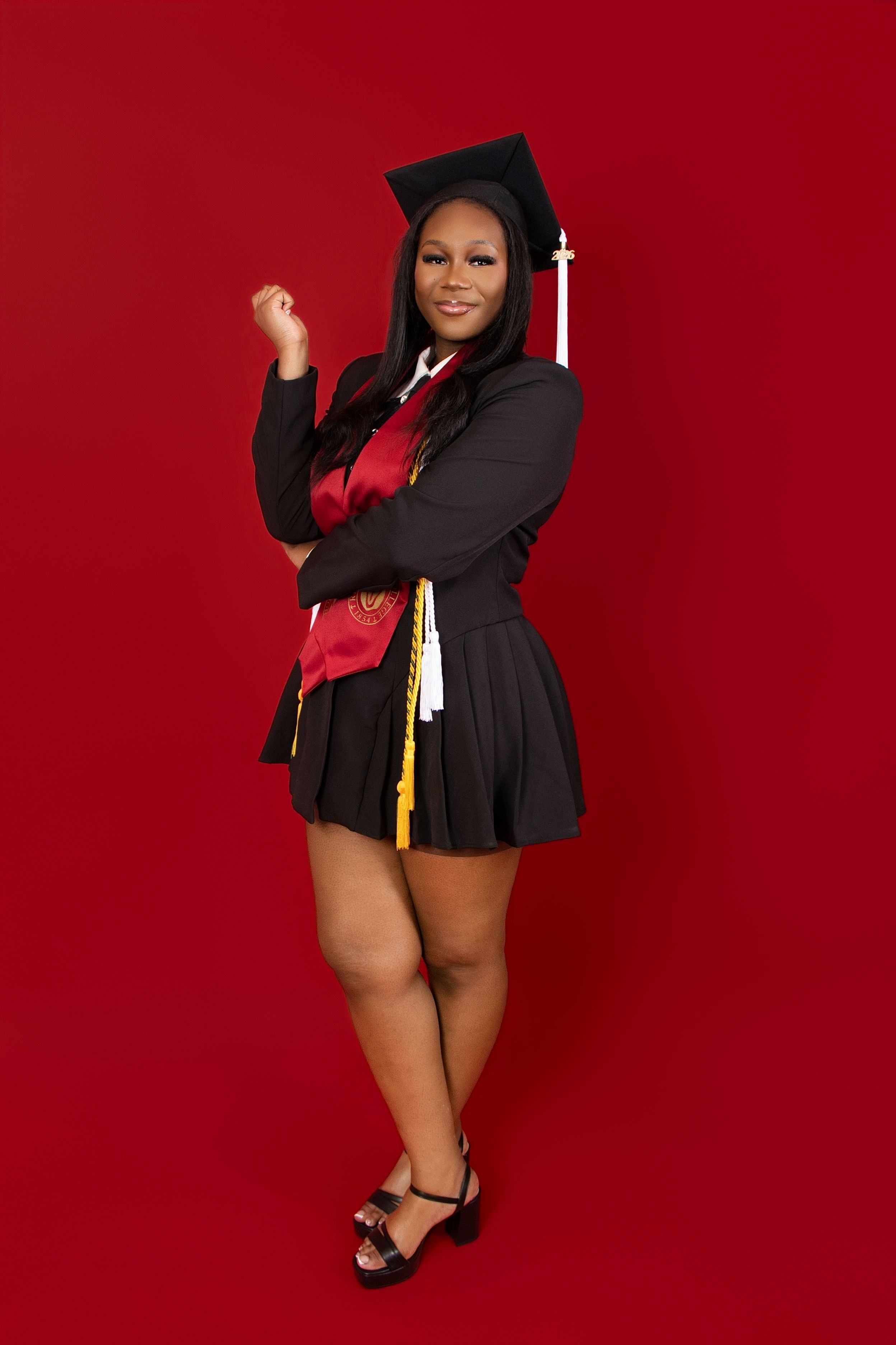 A young woman in black graduation cap and gown with red sash poses confidently against a bold red background.