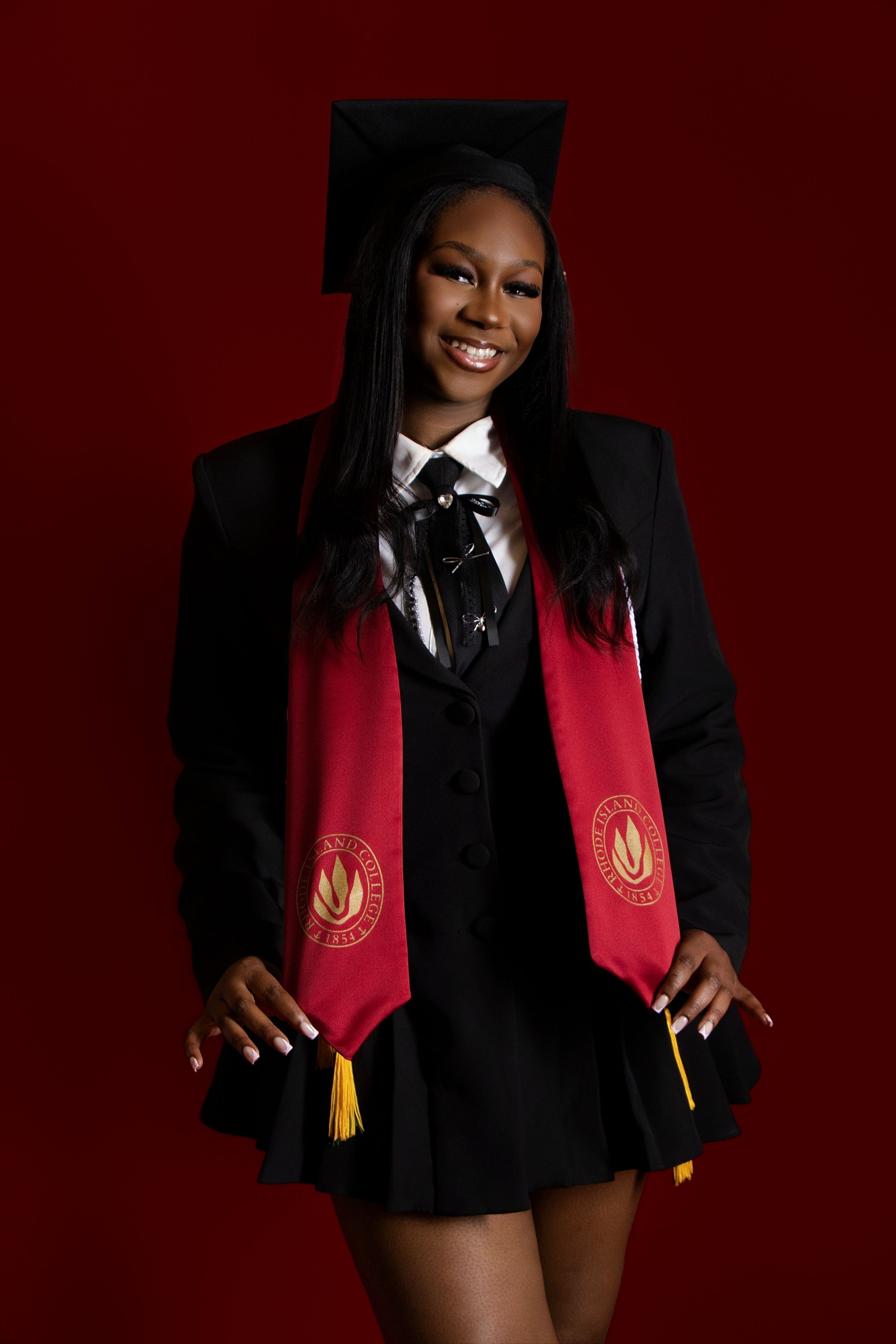 Smiling graduate in black cap, gown, and red stole poses confidently against a dark red background.