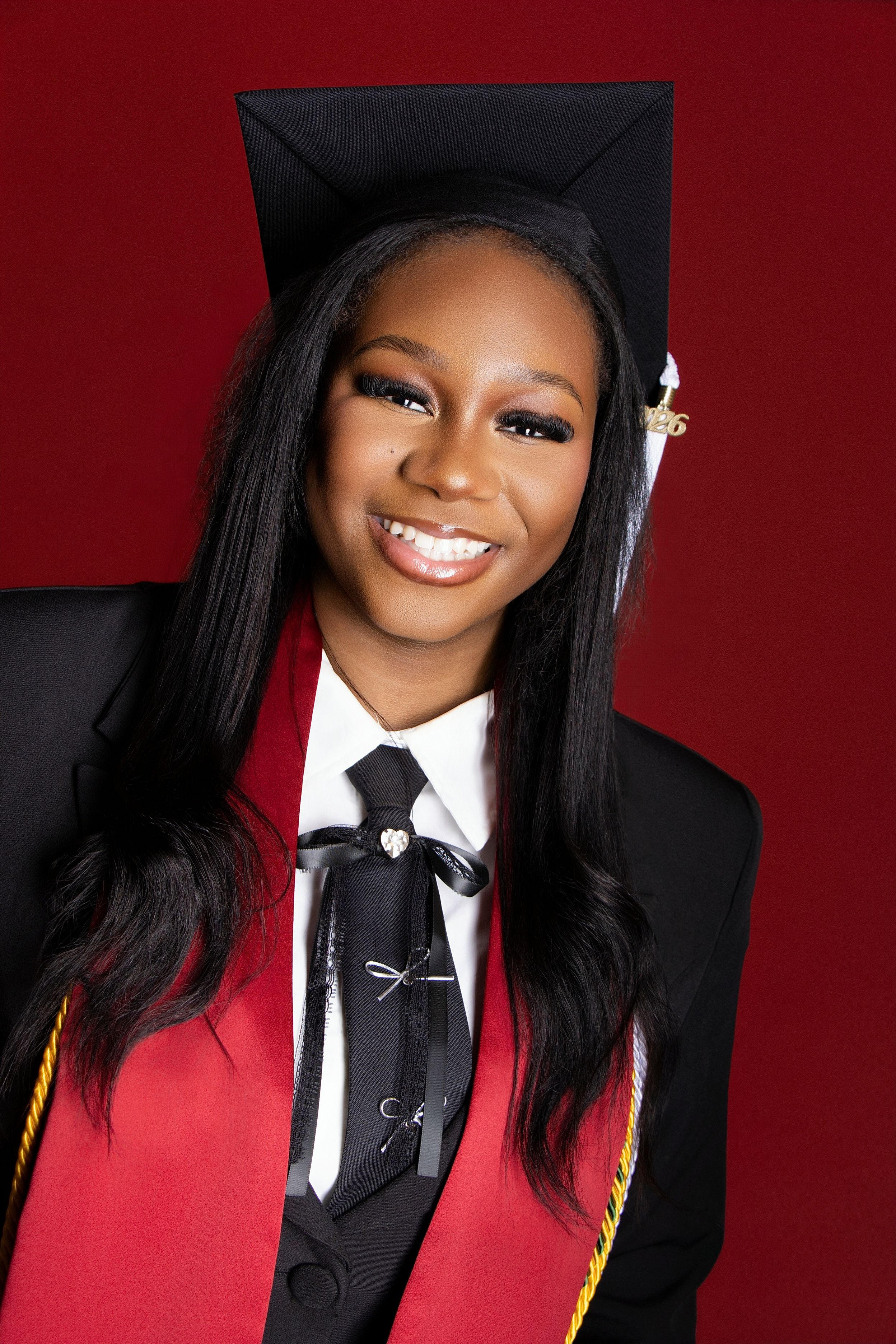 Smiling young woman in red graduation gown and black cap with gold honor cords poses against red background.