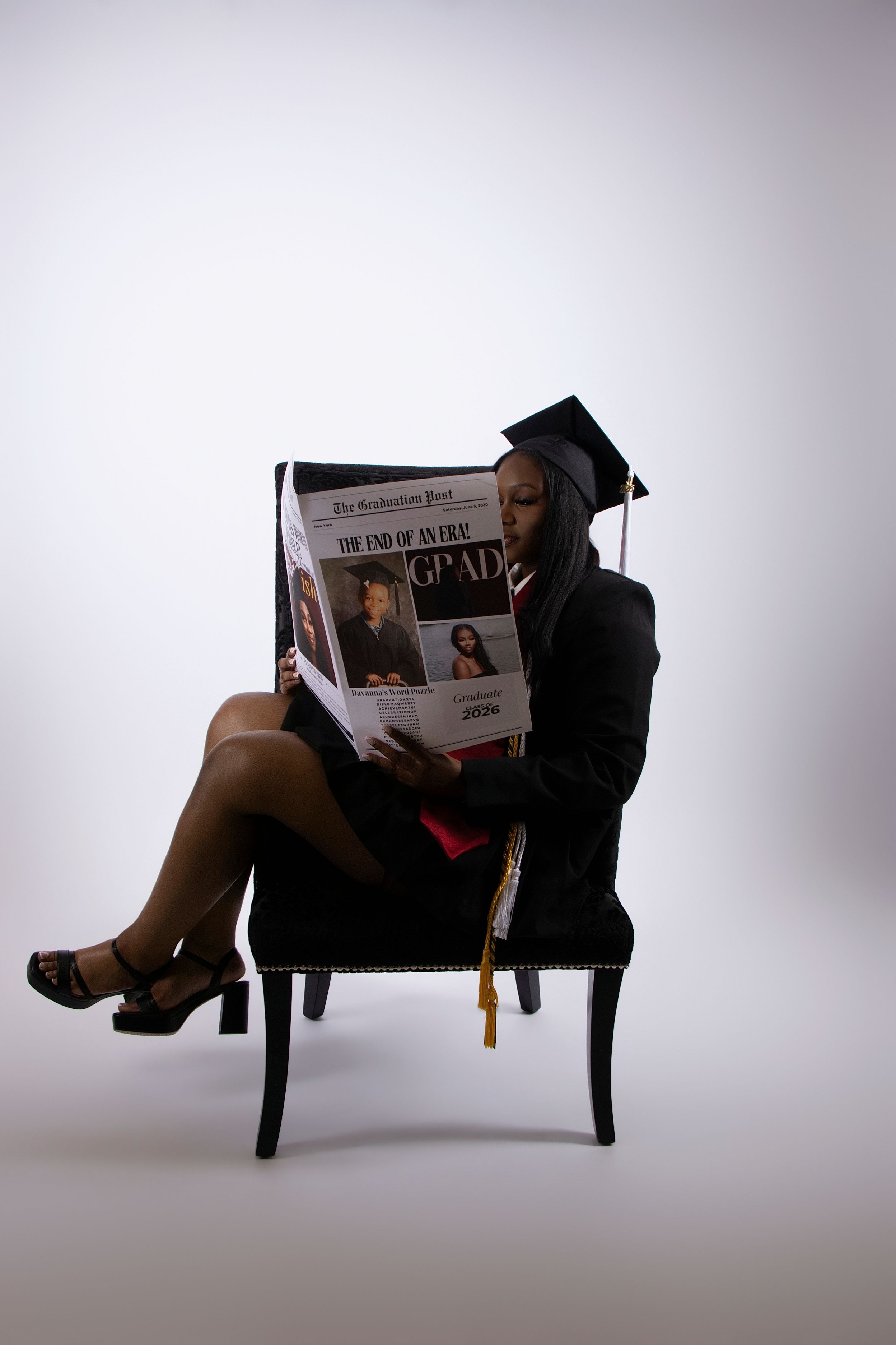 Graduate in cap and gown sits in chair reading a Washington Post newspaper with GRAD headline.
